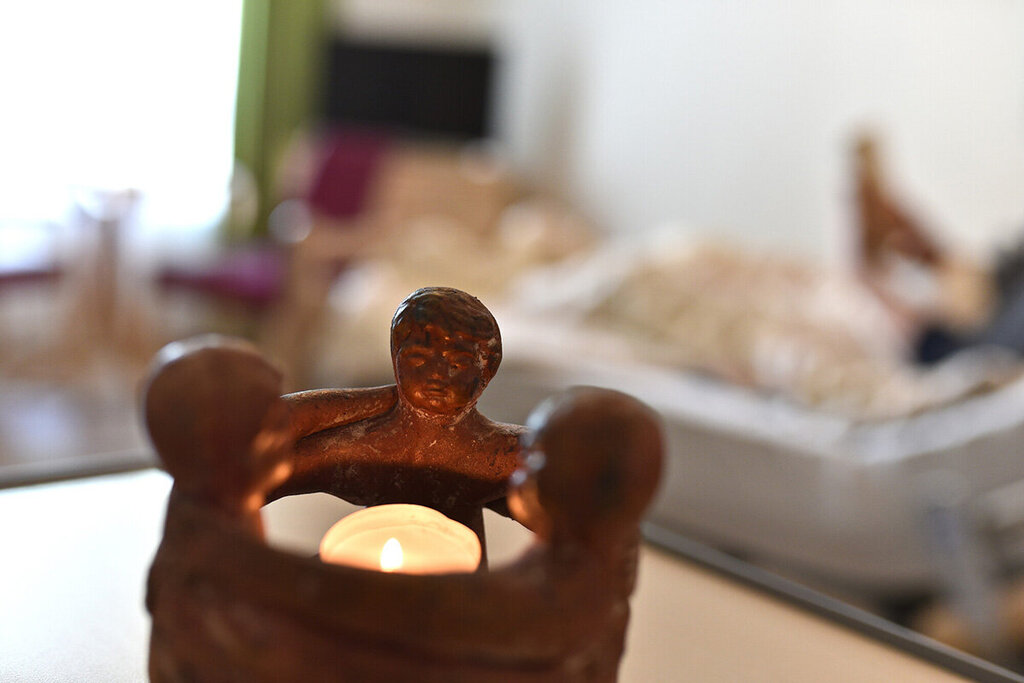 Im Hintergrund verschwommen liegt eine Person in einem Bett. Im Vordergrund ist ein Teelicht zu sehen, welches von drei Figuren umschlossen wird.