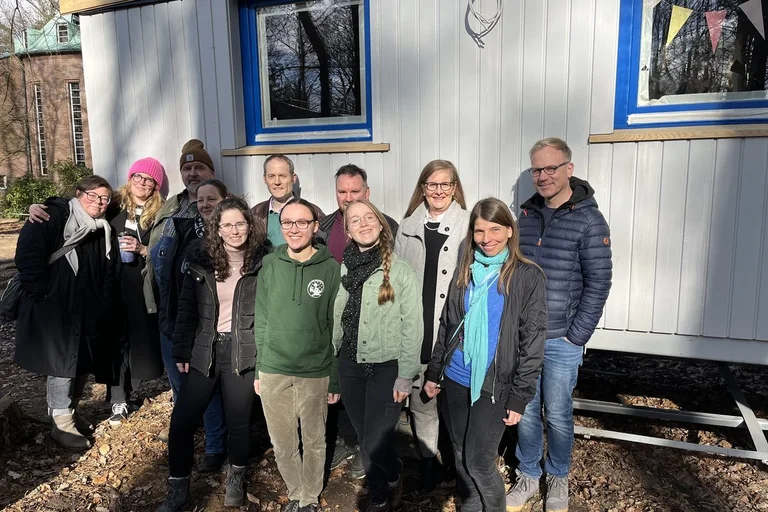 Eine Menschengruppe steht vor einem Bauwagen und lächelt.