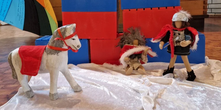 Spielfiguren: Ein Pferd mit roter Decke und rotem Zaunzeug, eine Figur mit Helm und Schwert steht und eine Figur mit rotem Tuch und weißem Peltz sitzt auf einer weißen Satindecke, im Hintergrund große rote und blaue Kästen