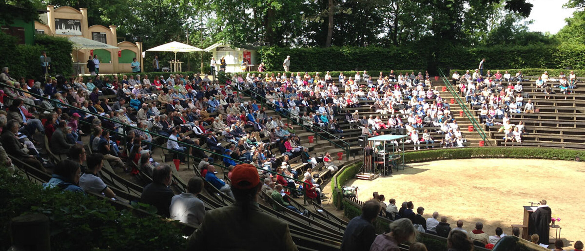 Gottesdienst im Freien - Im Halbkreis, mehrere hintereinander angeordnete Sitzreihen mit stizenden Menschen. DAs Ganze ist mit Bäumen umrahmt, die Sonne scheint, 2/3 der Leute sitzt im Schatten. 