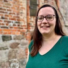 Junge Frau lächelt vor einer Backsteinkirche stehend in die Kamera.