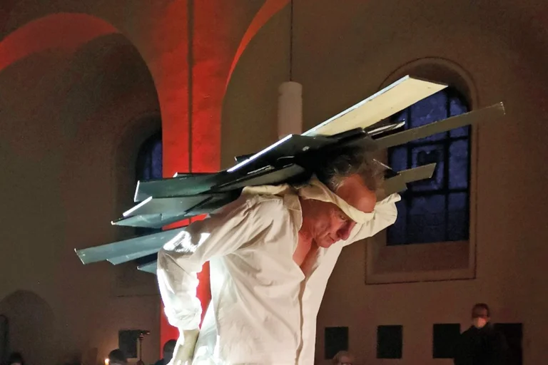 Ein Mann im weißen Hemd trägt etwas auf dem Rücken, dass an das Kreuz Jesu erinnert.