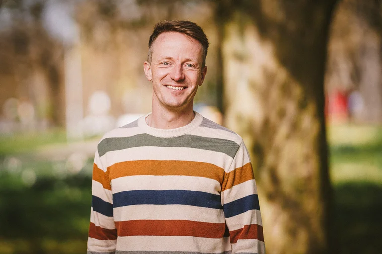 Mann steht vor einem Baum und lächelt in die Kamera.