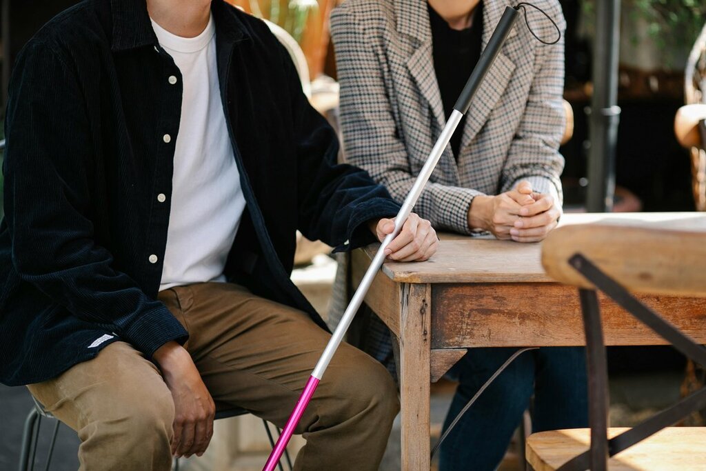Zwei Personen, deren Köpfe nicht zu sehen sind, sitzen an einem Tisch mit einem Blindenstock. 