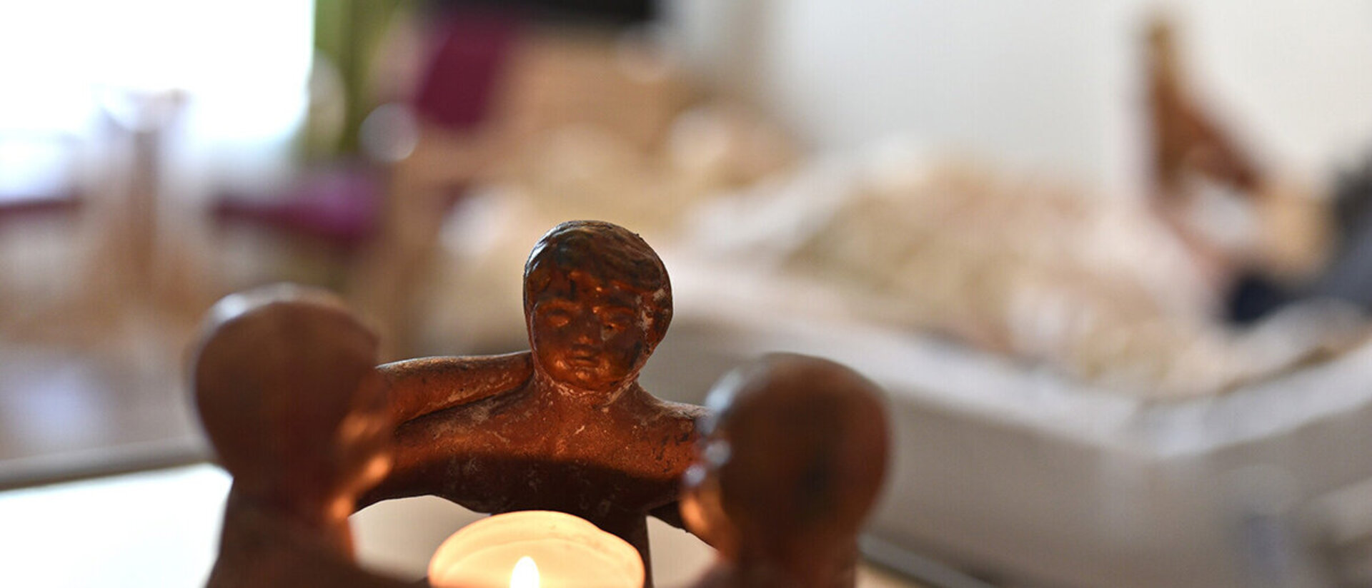 Im Hintergrund verschwommen liegt eine Person in einem Bett. Im Vordergrund ist ein Teelicht zu sehen, welches von drei Figuren umschlossen wird.