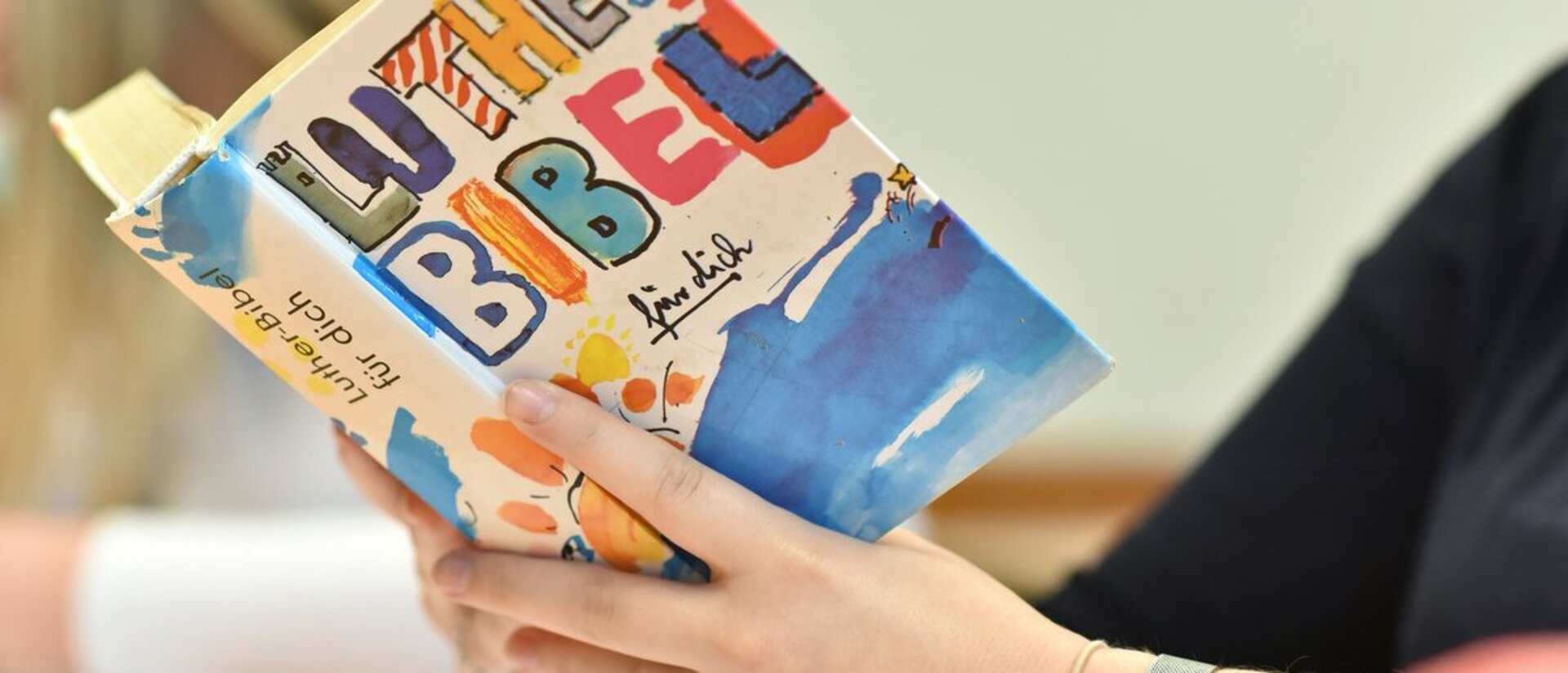 Hände halten eine aufgeschlagene bunt gestaltete Bibel