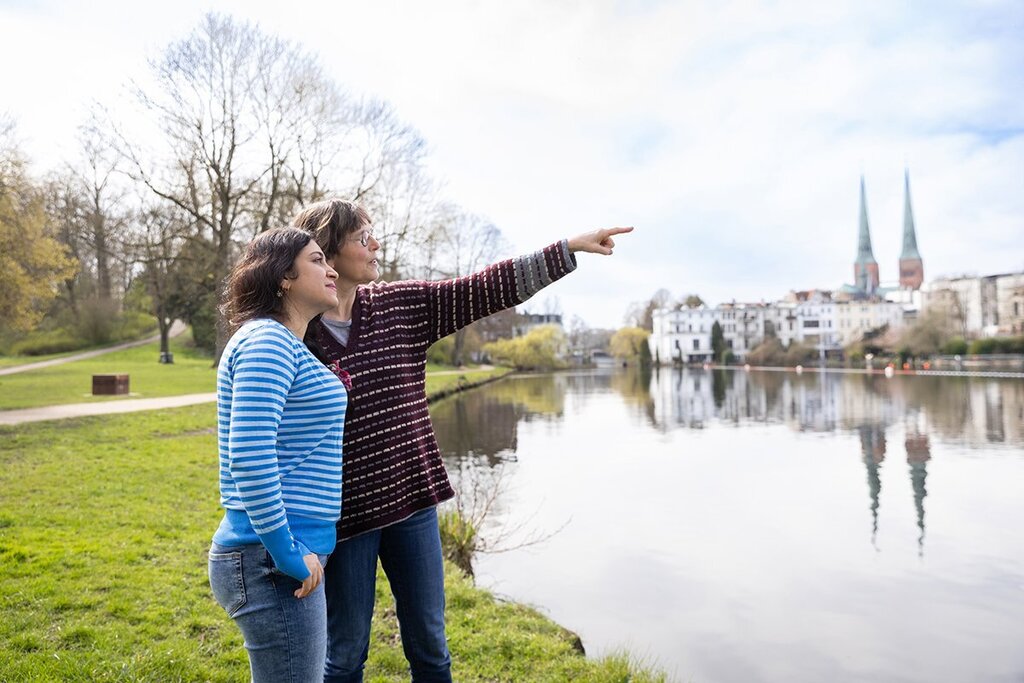 Zwei Frauen stehen nebeneinander an einem Gewässer. Die eine Frau zeigt in die Ferne. Im Hintergrund ist die Stadt Lübeck zu sehen mit den Türmen von St. Marien und dem Petriturm.