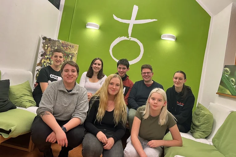 Eine Gruppe junger Menschen hockt vor einer grün gestrichenen Wand. 