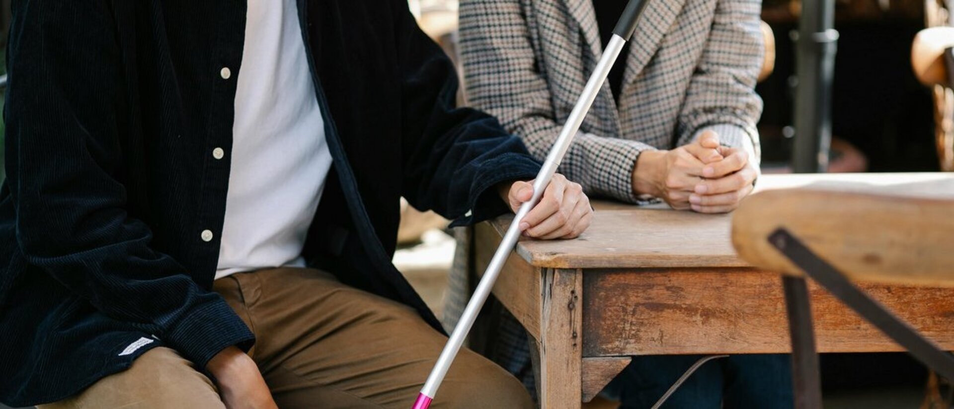 Zwei Personen, deren Köpfe nicht zu sehen sind, sitzen an einem Tisch mit einem Blindenstock. 