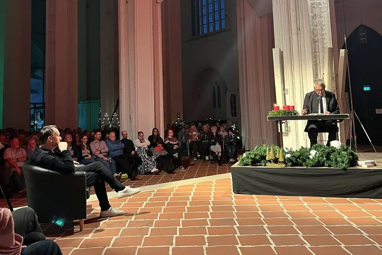 Ein Mann sitzt in einem Sessel und der andere am Tisch und liest mit Mikro