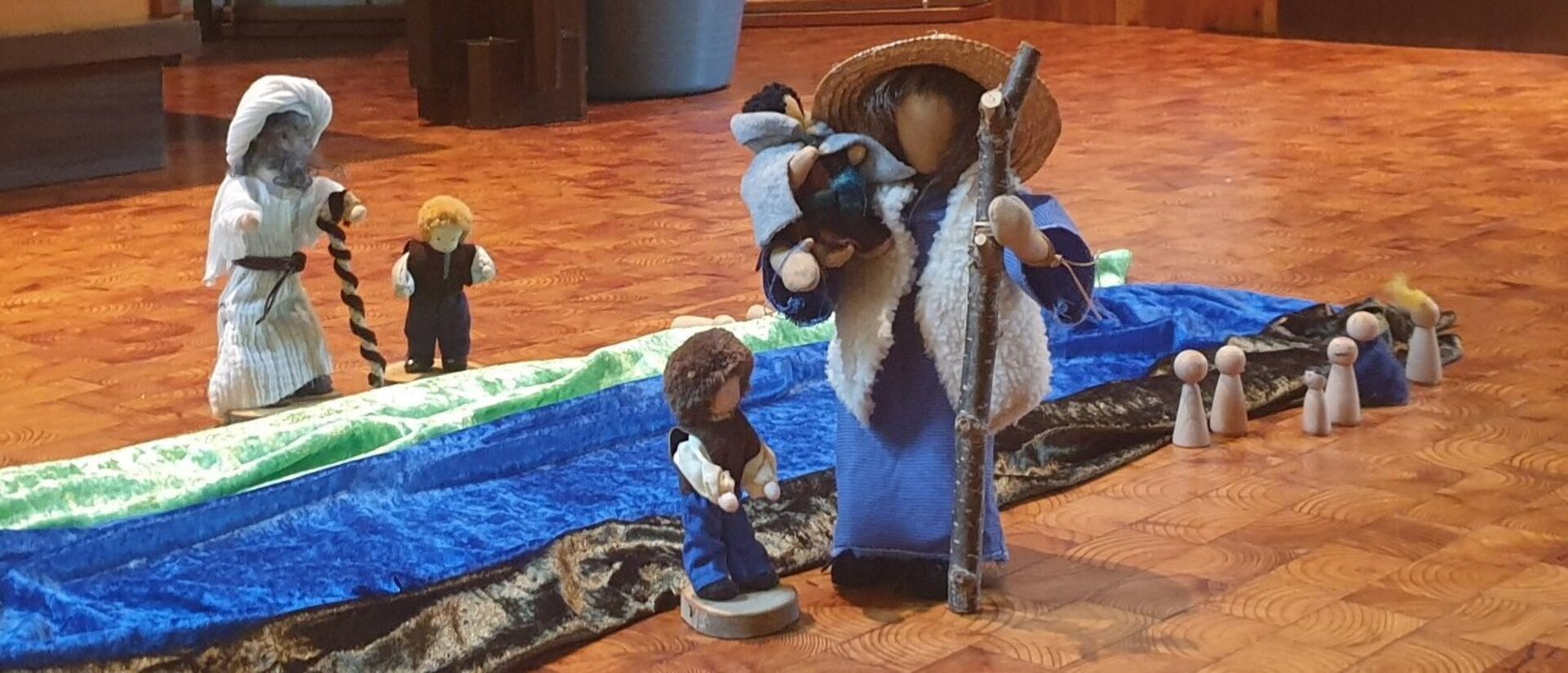 Mehrere Figuren auf einem Kirchenboden, kleine Kinder ein Erwachsener mit einem Kind auf dem Arm, mehrere Farbige Tücher liegen längs nebeneinander - Farben grün, blau und braun