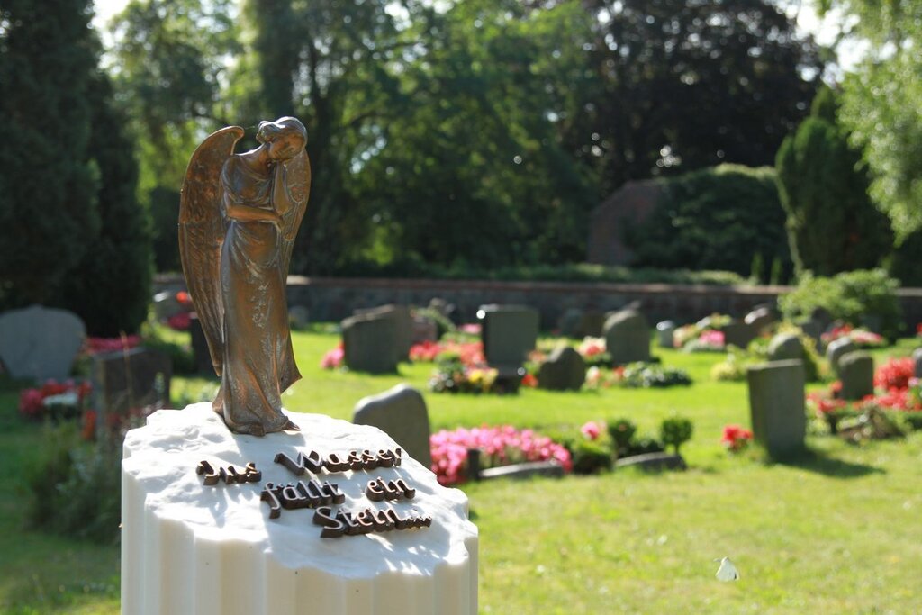 Ein Metallengel auf einem weißen sockel mit der Aufschrift: Ins Wasser fällt ein Stein - Im Hintergrund sind Grabsteine, Rasenfläche und Blumen zu sehen. Es scheint die Sonne