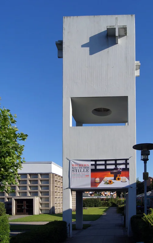 Beton-Glockenturm mit Spannband 'Kirche der Stille', darunter Schatten, links Ecke des Kirchsaals mit Eingang, am Rand grüne junge Linde, rechts eine Laterne
