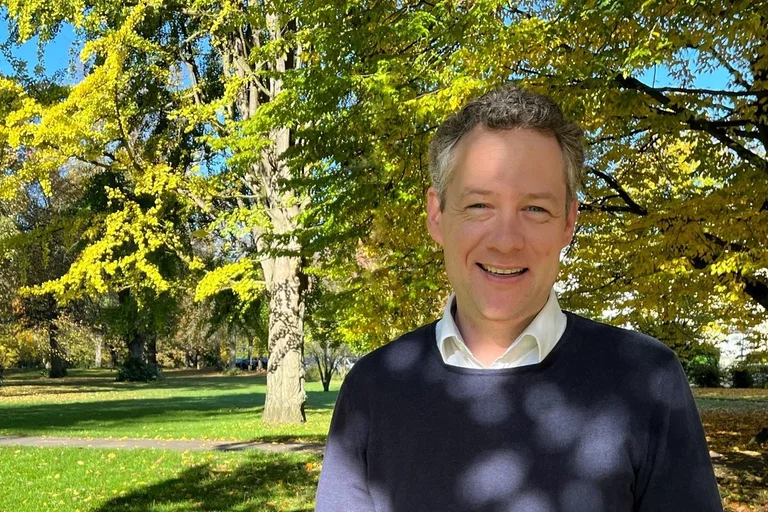 Mann steht vor einem Laubbaum und lächelt  in die Kamera.