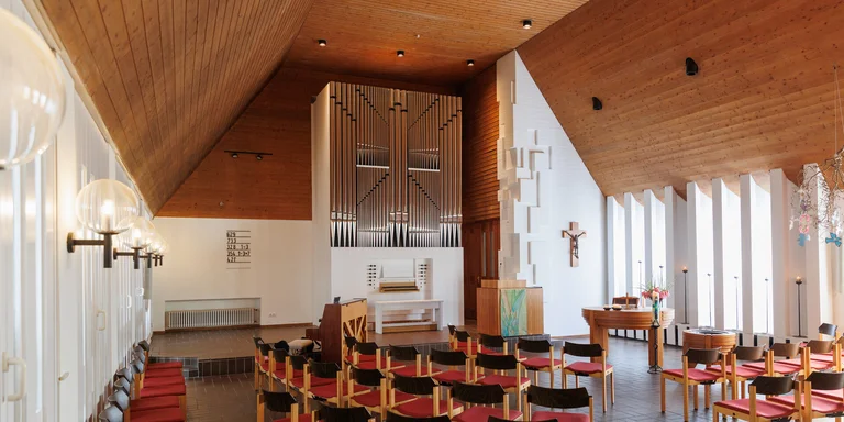 Innenraum einer Kirche, die Decke ist mit hellem Holz getäfelt man sieht eine Orgel mit Orgelpfeifen im Hintergrund, davor mehrere Stuhlreihen.