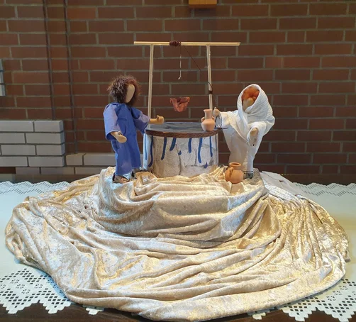 zwei Puppen stehen an einem gebstelten Brunnen, sie stehen auf einem beigen Satintuch 