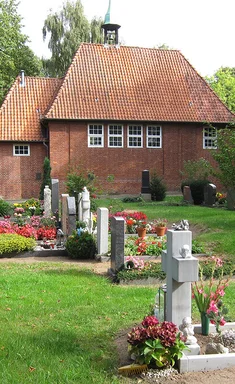 Vor der Friedhofskapelle sind mehrere Gräber, mit blühenden Pflanzen und ein Weg zu sehen