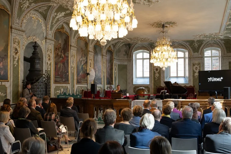 Eine Reihe von Menschen sitzt in einem barocken festlichem Saal, zwei Kronleuchter hängen von der Decke