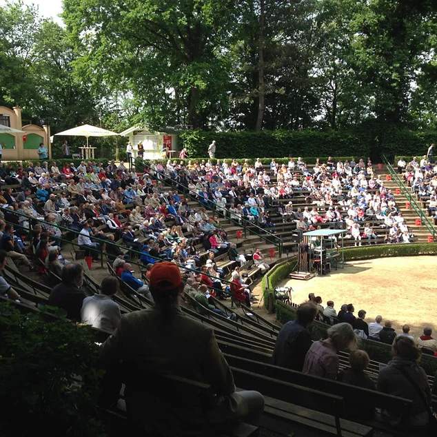 Gottesdienst im Freien - Im Halbkreis, mehrere hintereinander angeordnete Sitzreihen mit stizenden Menschen. DAs Ganze ist mit Bäumen umrahmt, die Sonne scheint, 2/3 der Leute sitzt im Schatten. 
