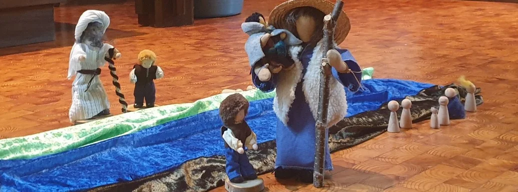 Mehrere Figuren auf einem Kirchenboden, kleine Kinder ein Erwachsener mit einem Kind auf dem Arm, mehrere Farbige Tücher liegen längs nebeneinander - Farben grün, blau und braun