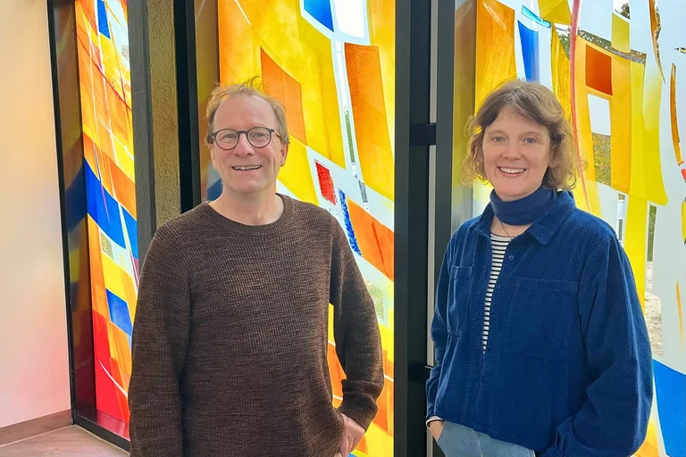 Ein Mann und eine Frau stehen nebeneinander vor den bunten Fenstern des "Raums der Stille".