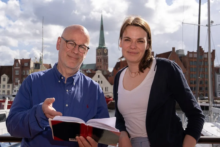 Zwei Menschen stehen an einem Gewässer und schauen in die Kamera