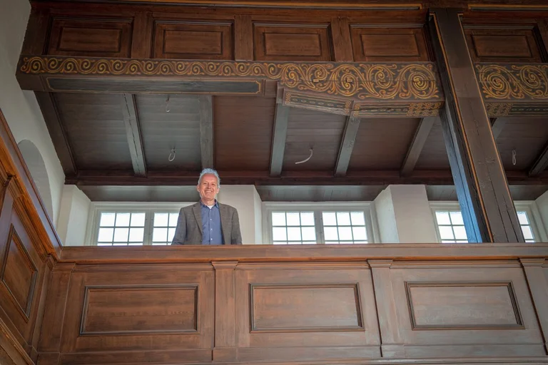 Ein Mann steht auf der hölzernen Empore einer Kirche und lächelt in die Kamera.