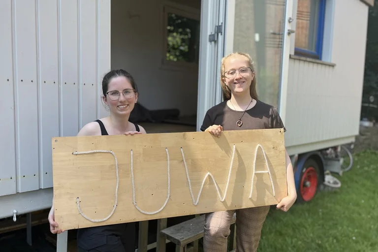 Zwei junge Frauen stehen vor einem Bauwagen und halten ein Schild mit der Aufschrift "JuWa".