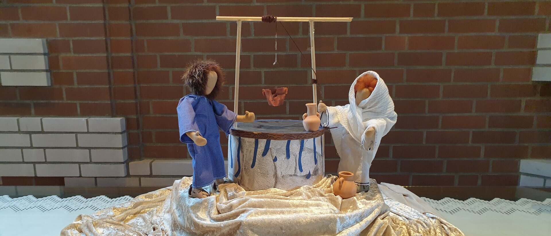 zwei Puppen stehen an einem gebstelten Brunnen, sie stehen auf einem beigen Satintuch 